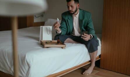 elegantly dressed man sitting on a bed and holding a cosmetic product